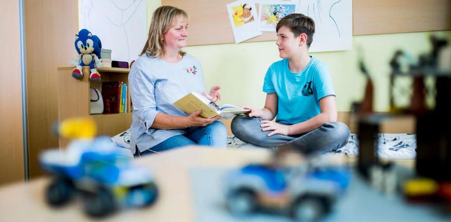 Eine Therapeutin liest einem jungen Pateinten der Jugendpsychiatrie in seinem Patientenzimmer aus einem Buch vor