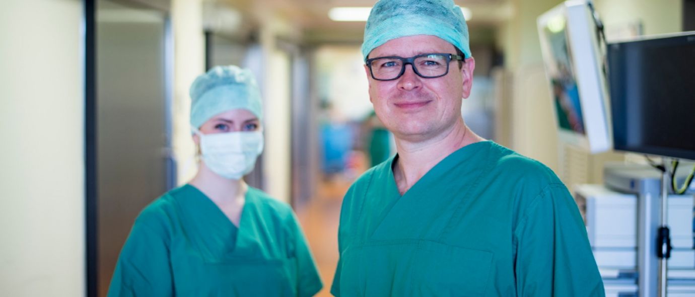 Arzt und eine Op-Pflegerin mit Mundschutz blicken in die Kamera
