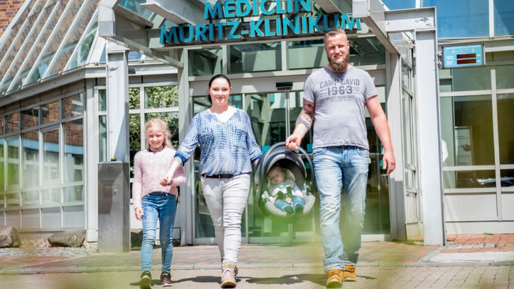 Eine junge Familie mit einer Tochter und einem Neugeborernen verlässt das MEDICLIN Müritz-Klinikum durch den Haupteingang