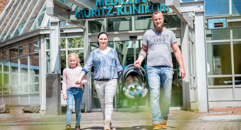 Eine junge Familie mit einer Tochter und einem Neugeborernen verlässt das MEDICLIN Müritz-Klinikum durch den Haupteingang