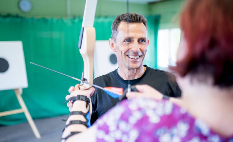 Patientin und Therapeut beim Bogenschießen in der Sporthalle